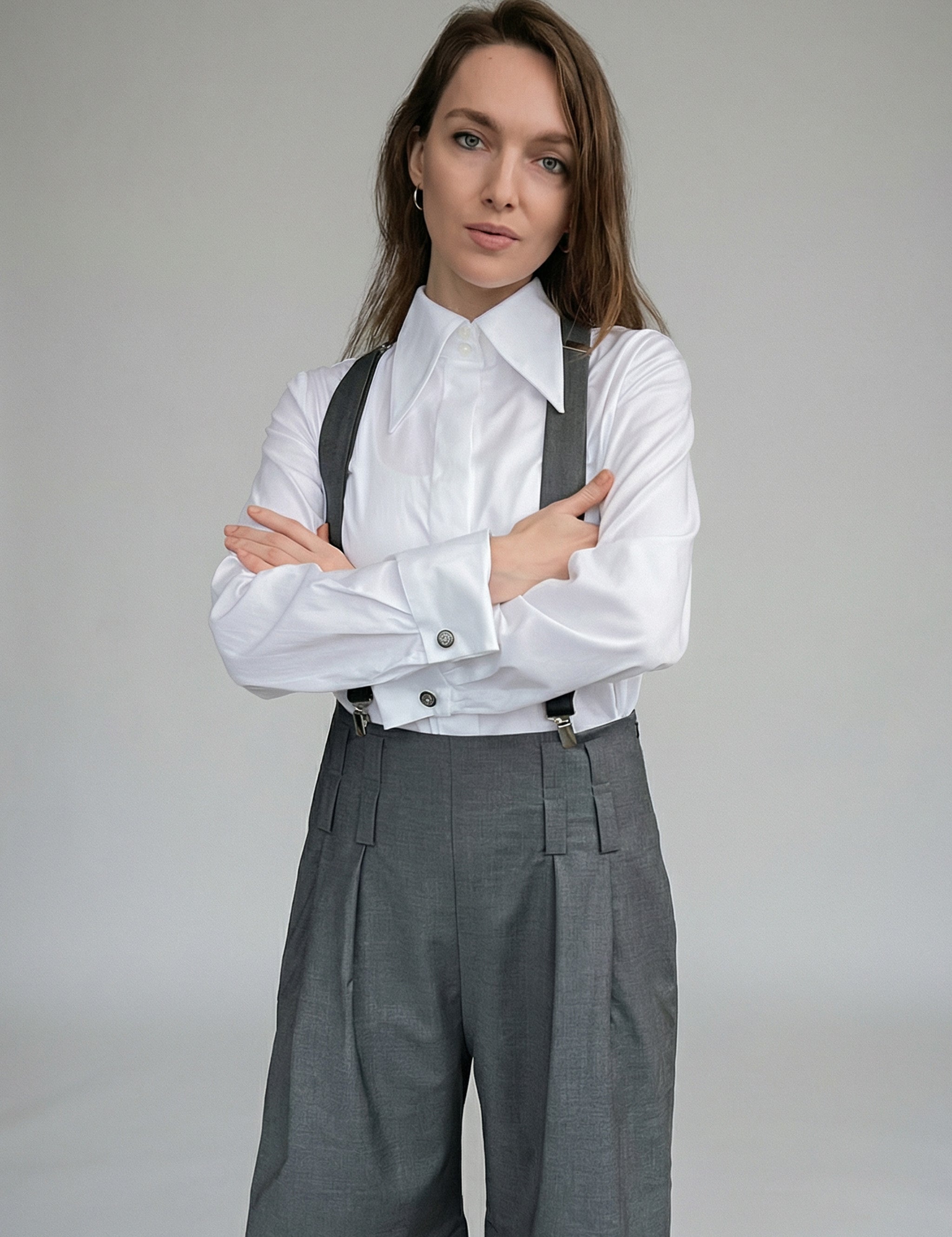 Woman wearing a white shirt with rolled-up sleeves and gray suspenders over gray pants on a plain background