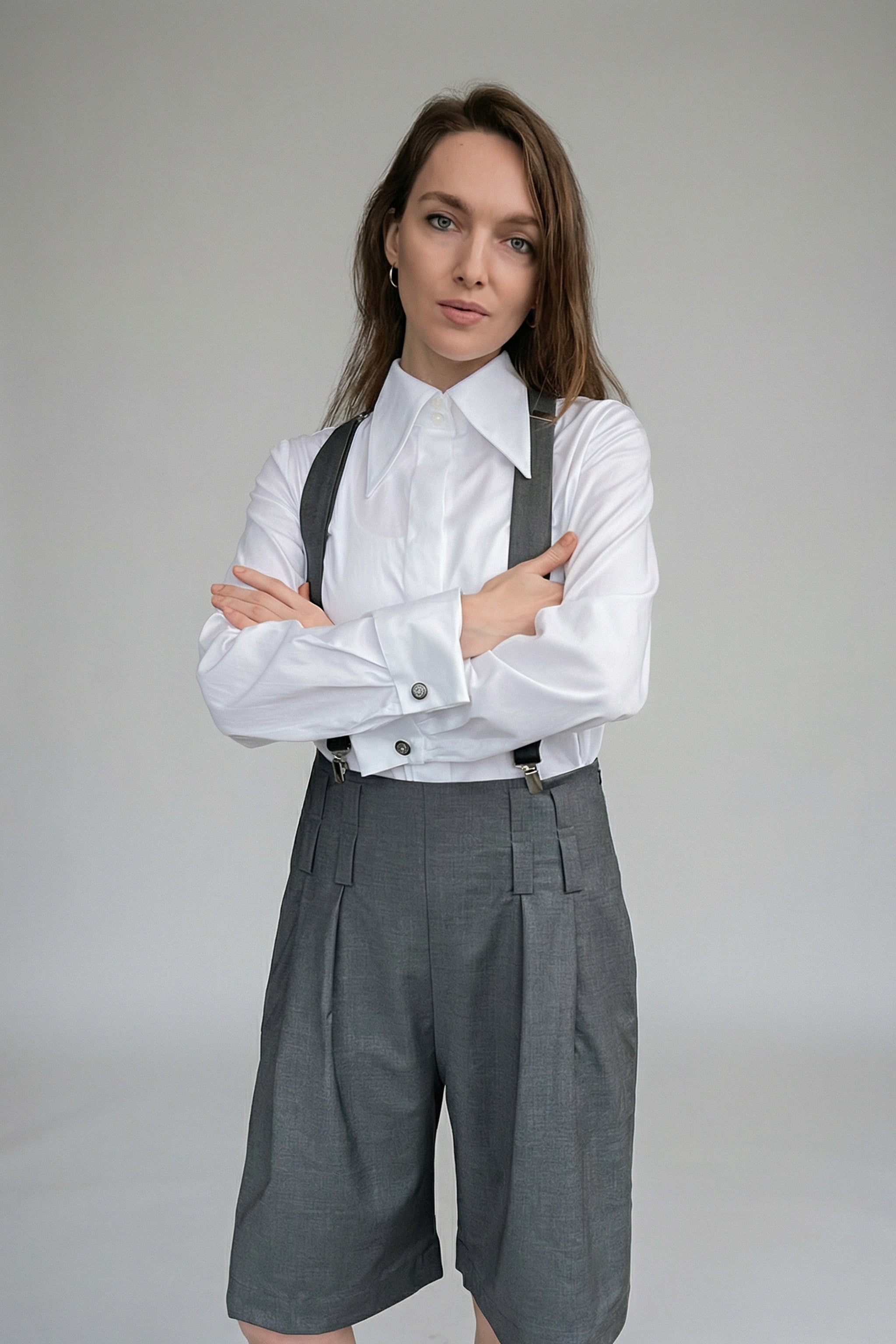 Woman wearing a white shirt with rolled-up sleeves and gray suspenders over gray pants on a plain background