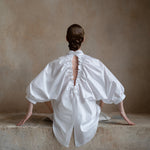 Full-length view of a seated model with arms outstretched, showcasing a white open-back shirt and puff sleeves