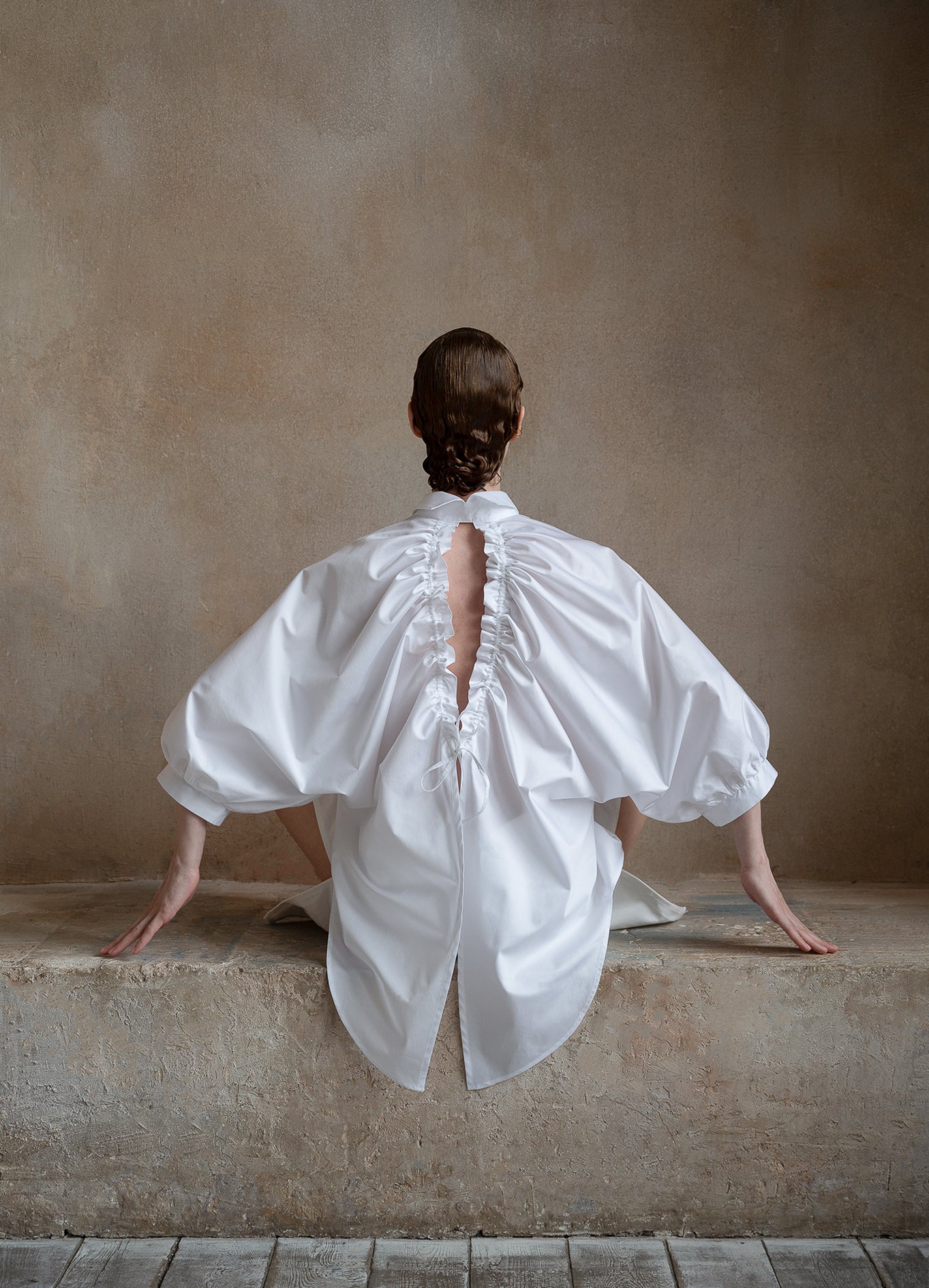 Full-length view of a seated model with arms outstretched, showcasing a white open-back shirt and puff sleeves