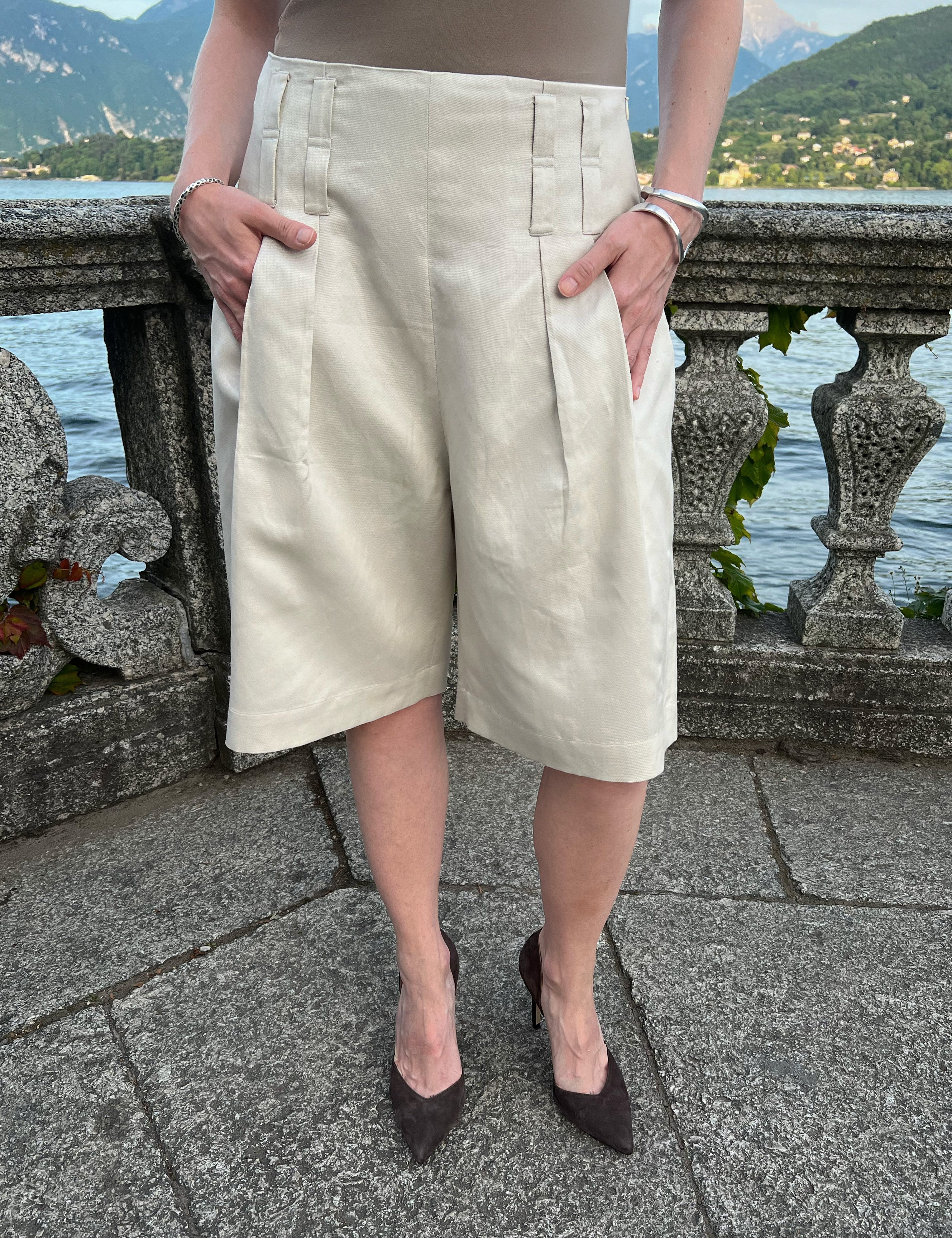 Sand linen-blend Bermuda shorts with front pleats and double belt loops, worn by a model at Lake Como, Italy