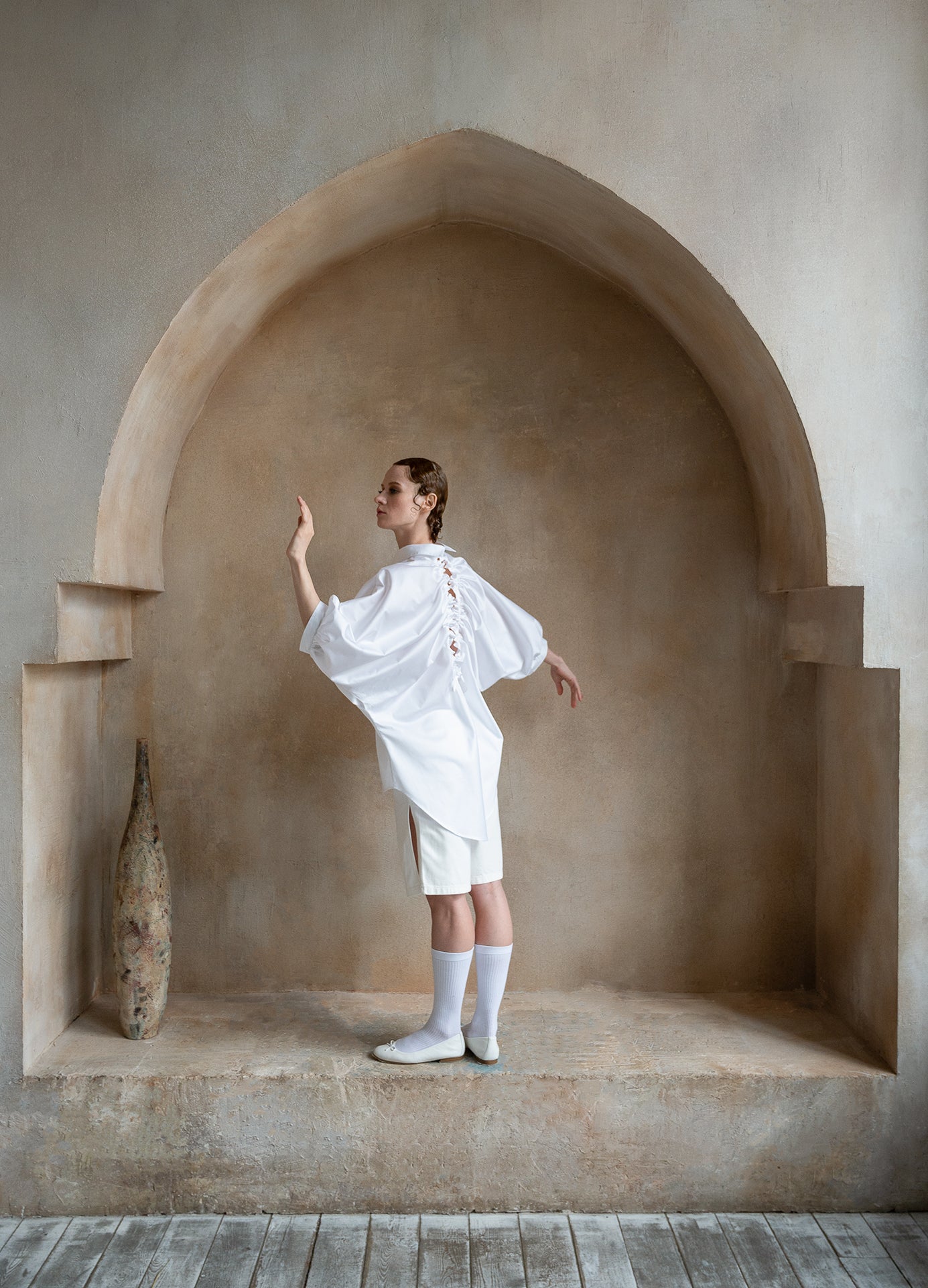White open-back shirt worn by a model beneath a sand-toned arch, showcasing the signature drawstring back