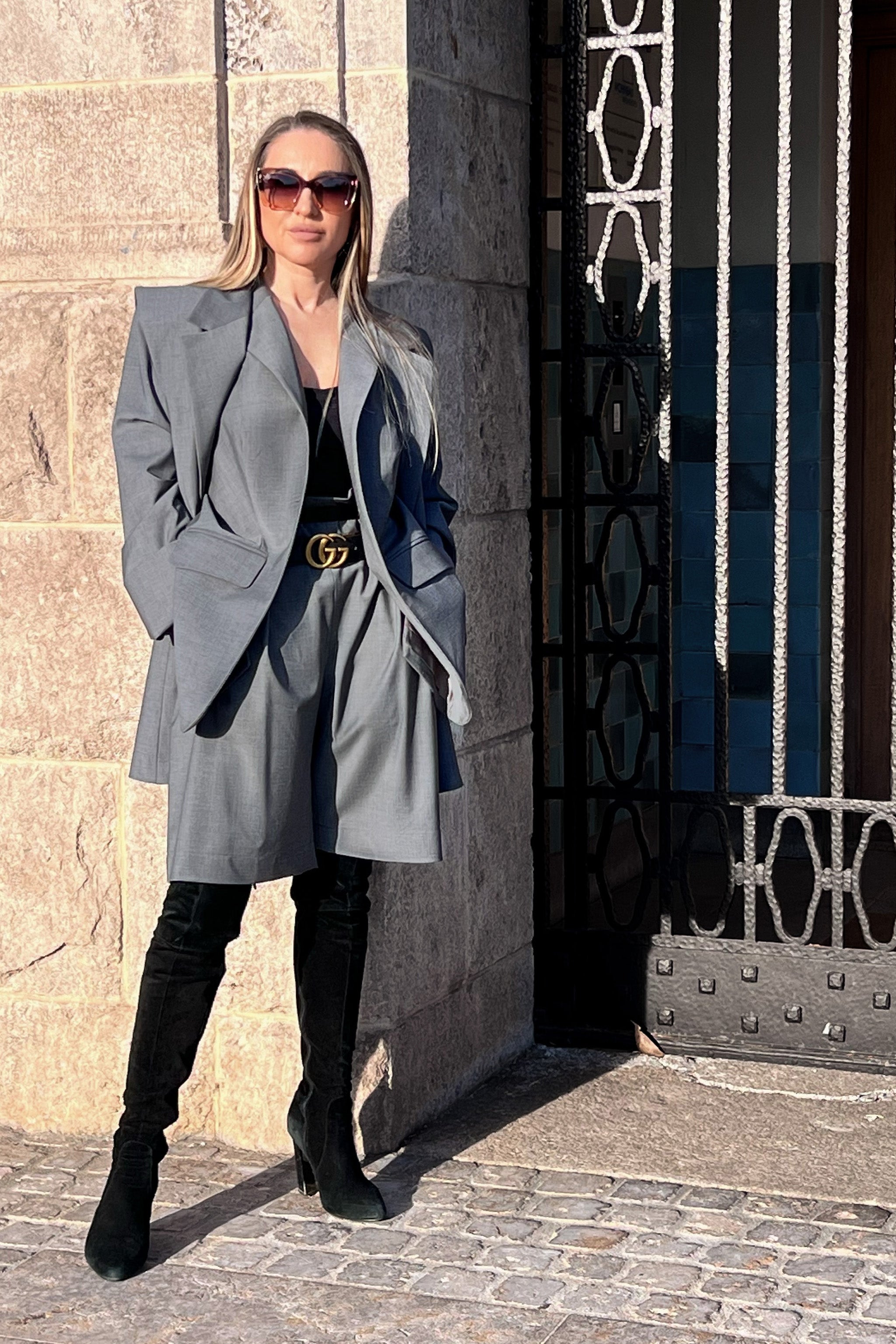 Woman in a gray suit and black boots standing against a stone wall