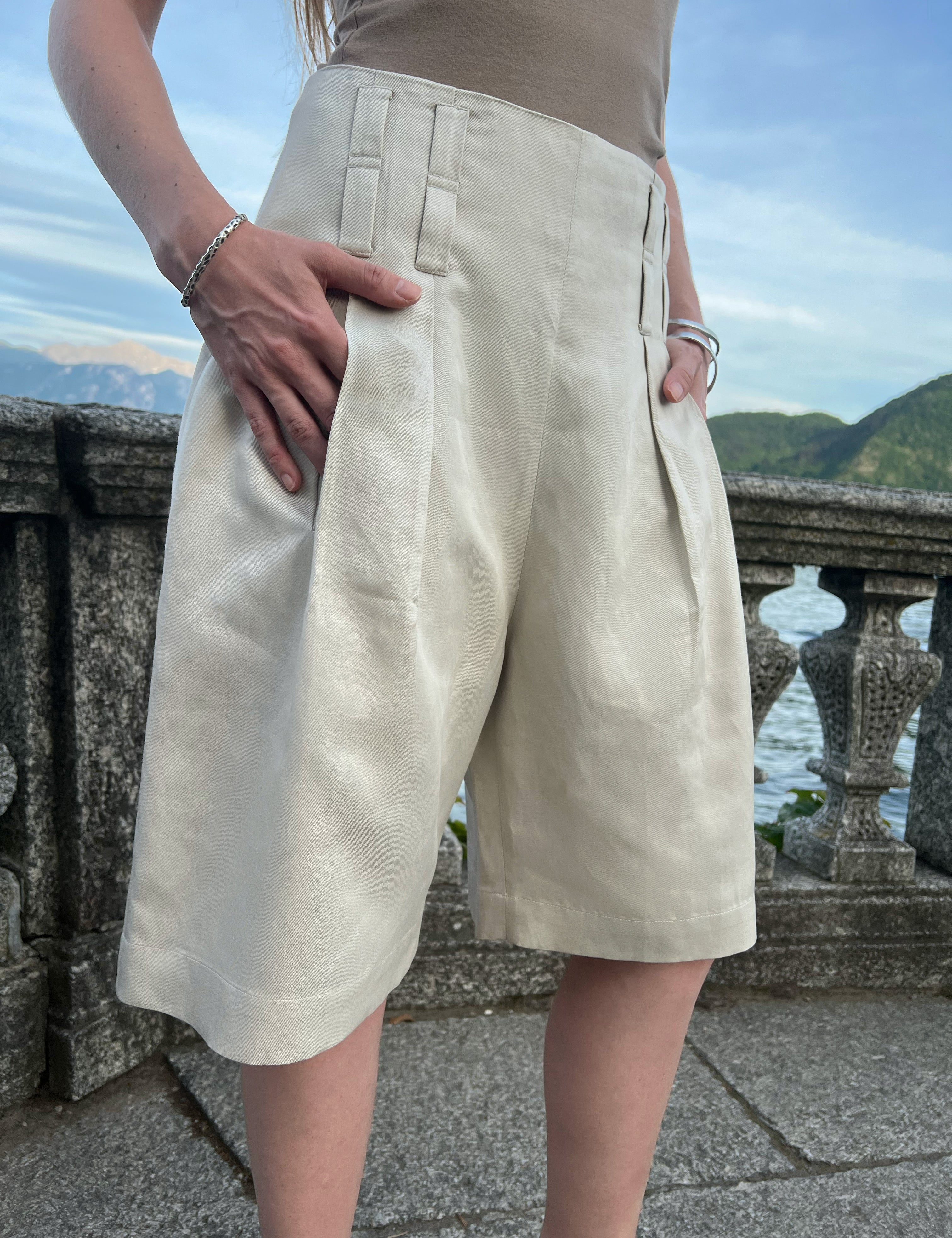 Front view of pleated sand Bermuda shorts featuring knee length, refined tailoring and a relaxed silhouette.